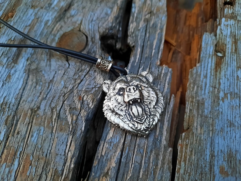May include: A silver bear head pendant necklace with a black leather cord. The bear's mouth is open and its teeth are visible.