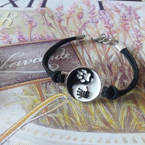 May include: Black bracelet with a silver yin yang charm featuring paw prints. The charm is black and white with a silver outline.