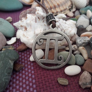 Peut inclure: Un pendentif en argent représentant le signe du zodiaque des Gémeaux sur un cordon en cuir brun. Le pendentif est entouré de pierres lisses grises et brunes.