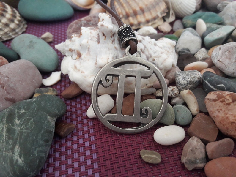 May include: A silver Gemini zodiac sign pendant on a brown leather cord. The pendant is surrounded by smooth, gray and brown stones.