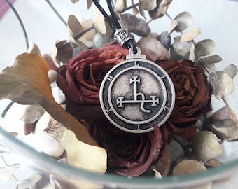 May include: A silver pendant with a sigil design, featuring a cross and a symbol, surrounded by the letters 'L', 'I', 'H', 'I', 'T', and 'L'. The pendant is resting on dried roses and leaves in a glass bowl.