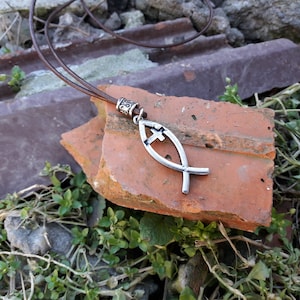 Puede incluir: Un collar de cordón de cuero marrón con un colgante de plata en forma de pez con una cruz en su interior. El collar está sobre un ladrillo rojo roto.