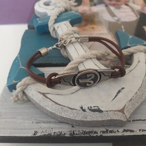 May include: A brown leather bracelet with a silver charm featuring the astrological symbol for Aries. The bracelet is wrapped around a white wooden anchor with blue accents.