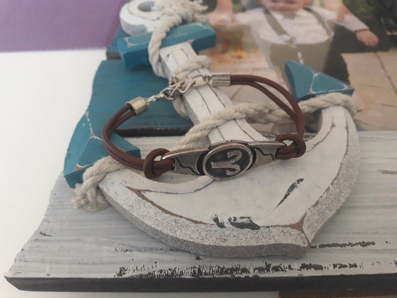 May include: A brown leather bracelet with a silver charm featuring the astrological symbol for Aries. The bracelet is wrapped around a white wooden anchor with blue accents.