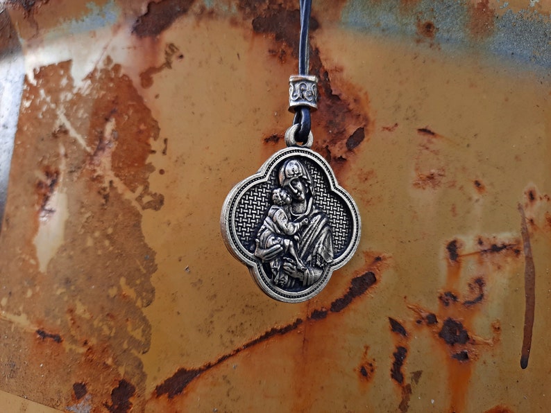 May include: A silver pendant necklace with a religious image of a mother and child. The pendant is in the shape of a four-leaf clover and has a woven pattern.