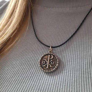 May include: A silver pendant necklace with a round pendant featuring the Chi Rho symbol, a Christian symbol that represents Christ. The pendant is surrounded by a circle of small beads. The necklace is on a black cord.
