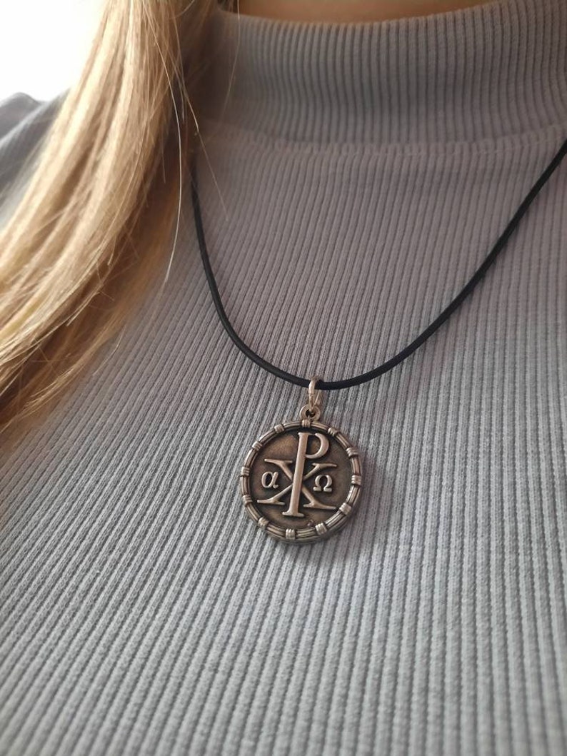 May include: A silver pendant necklace with a round pendant featuring the Chi Rho symbol, a Christian symbol that represents Christ. The pendant is surrounded by a circle of small beads. The necklace is on a black cord.