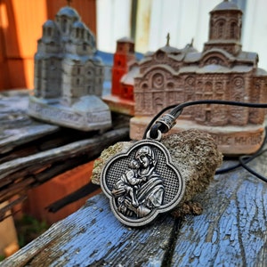May include: A silver pendant necklace with a religious image of the Virgin Mary holding a baby Jesus. The pendant is in the shape of a four-leaf clover and has a braided design around the edge. The necklace is lying on a wooden surface with small model churches in the background.