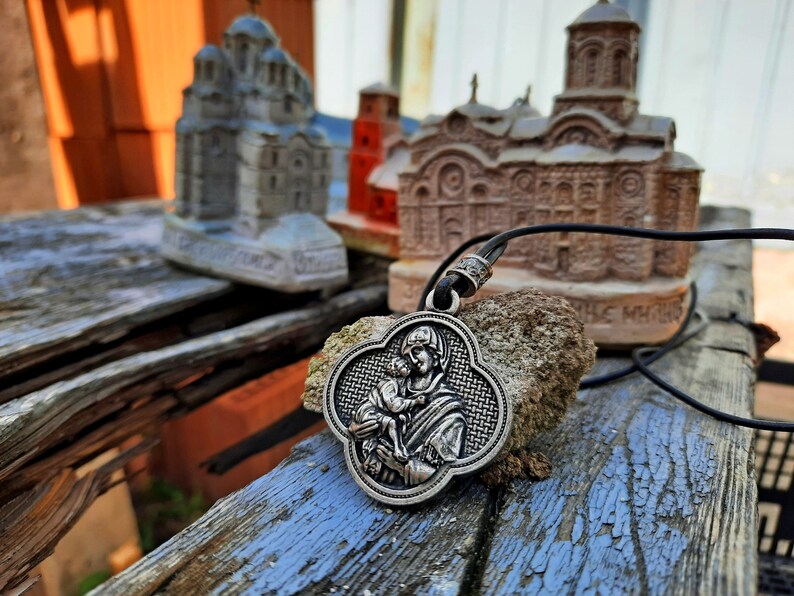 May include: A silver pendant necklace with a religious image of the Virgin Mary holding a baby Jesus. The pendant is in the shape of a four-leaf clover and has a braided design around the edge. The necklace is lying on a wooden surface with small model churches in the background.