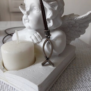 Puede incluir: Una figurita de ángel de cerámica blanca con alas, sosteniendo un cordón de cuero marrón con un colgante de Ichthys plateado. El ángel está sentado sobre una base blanca en forma de libro con una vela blanca en el centro.