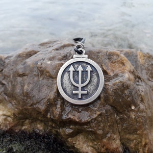 May include: A silver pendant necklace with a circular design featuring a trident and a symbol. The pendant is on a dark gray rock.