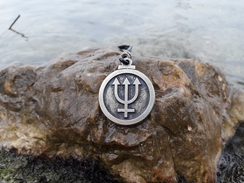 May include: A silver pendant necklace with a circular design featuring a trident and a symbol. The pendant is on a dark gray rock.
