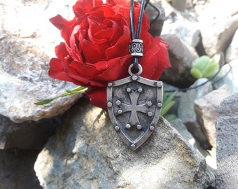 May include: A silver shield pendant with a cross design hangs from a black leather cord. The pendant is surrounded by a red rose.