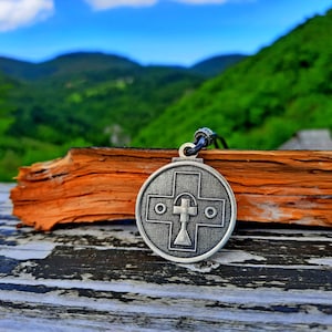 May include: A silver pendant necklace with a circular design featuring a cross inside. The pendant is on a wooden surface with a blurred background of green hills and a blue sky.