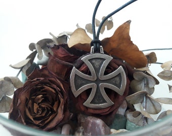May include: A silver cross pendant necklace with a black leather cord. The pendant is a Maltese cross with a textured surface. The necklace is displayed on a bed of dried roses and eucalyptus leaves.