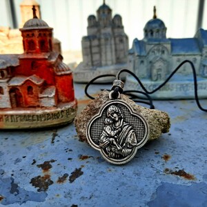 May include: A silver pendant necklace with a detailed image of the Virgin Mary holding a baby Jesus. The pendant is shaped like a four-leaf clover and is hanging on a black cord. The necklace is resting on a piece of driftwood in front of miniature church models.