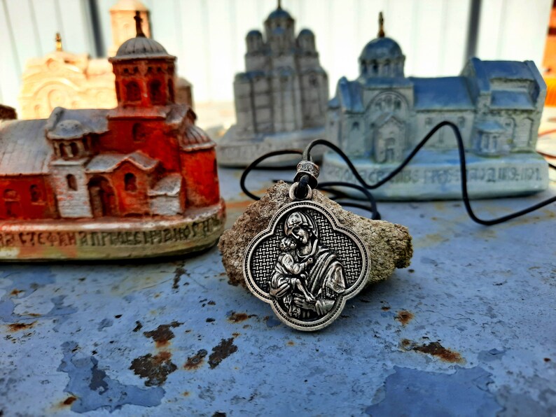 May include: A silver pendant necklace with a detailed image of the Virgin Mary holding a baby Jesus. The pendant is shaped like a four-leaf clover and is hanging on a black cord. The necklace is resting on a piece of driftwood in front of miniature church models.