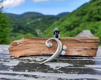 Puede incluir: Un colgante de clave de fa en plata sobre un cordón de cuero negro. El colgante está sobre una pieza de madera desgastada.