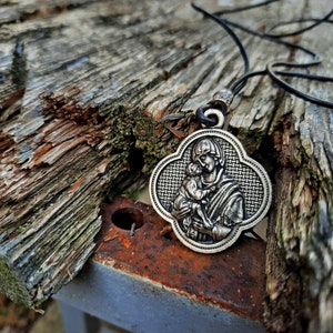 May include: A silver pendant necklace with a detailed image of a mother and child. The pendant is shaped like a four-leaf clover and has a black leather cord.