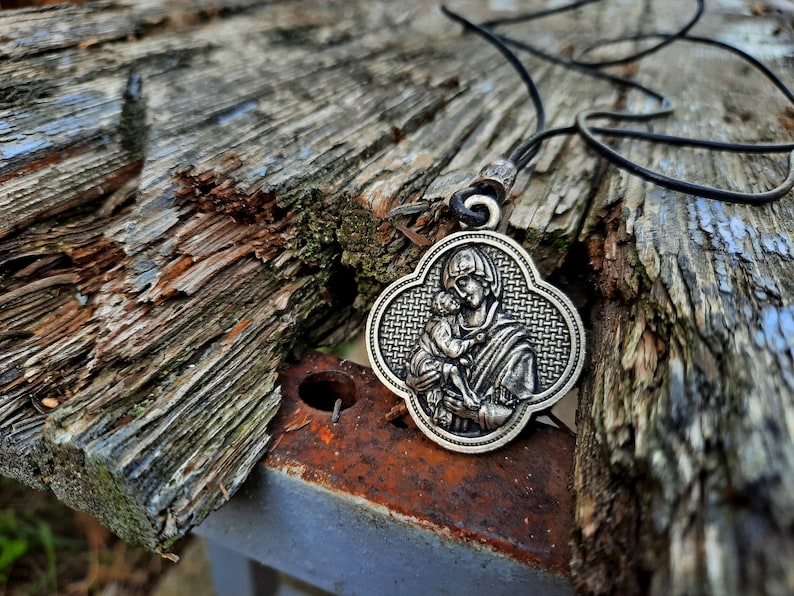 May include: A silver pendant necklace with a detailed image of a mother and child. The pendant is shaped like a four-leaf clover and has a black leather cord.