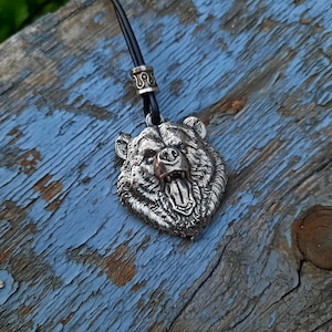 May include: A silver bear head pendant with a detailed, snarling expression, hanging from a black leather cord with a silver bead.