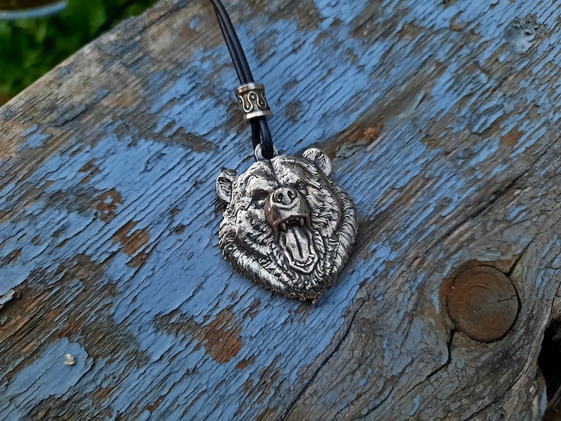 May include: A silver bear head pendant with a detailed, snarling expression, hanging from a black leather cord with a silver bead.