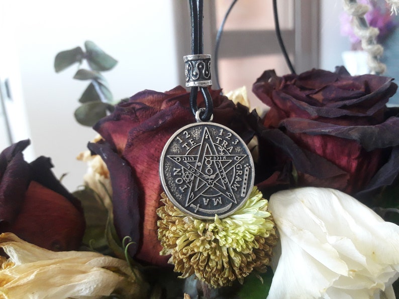 May include: A silver pendant necklace with a pentagram design and the words "1 2 3 TRA", "TE", "ON", "1 2 3", "GRA", "MA" around the edge. The necklace is hanging from a black leather cord and is displayed on a bouquet of dried roses.