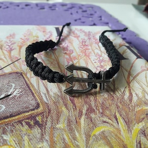 Może przedstawiać: Czarna pleciona bransoletka z srebrnym wisiorkiem w kształcie trójzębu. Bransoletka leży na powierzchni z kwiatowym wzorem.