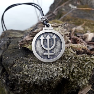 May include: A silver pendant necklace with a circular design featuring a trident and a symbol. The pendant is on a black leather cord and is resting on a piece of wood covered in moss.
