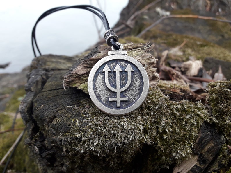 May include: A silver pendant necklace with a circular design featuring a trident and a symbol. The pendant is on a black leather cord and is resting on a piece of wood covered in moss.
