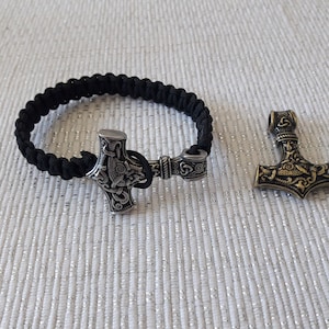 May include: A black paracord bracelet with a silver Mjolnir pendant. The pendant has intricate details and a braided design. A separate gold Mjolnir pendant is also shown.