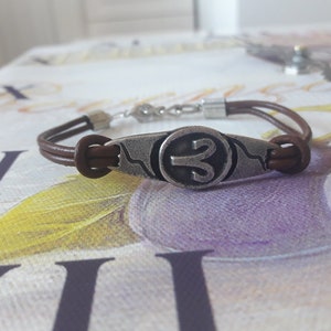 May include: A brown leather bracelet with a silver charm featuring the astrological symbol for Aries.