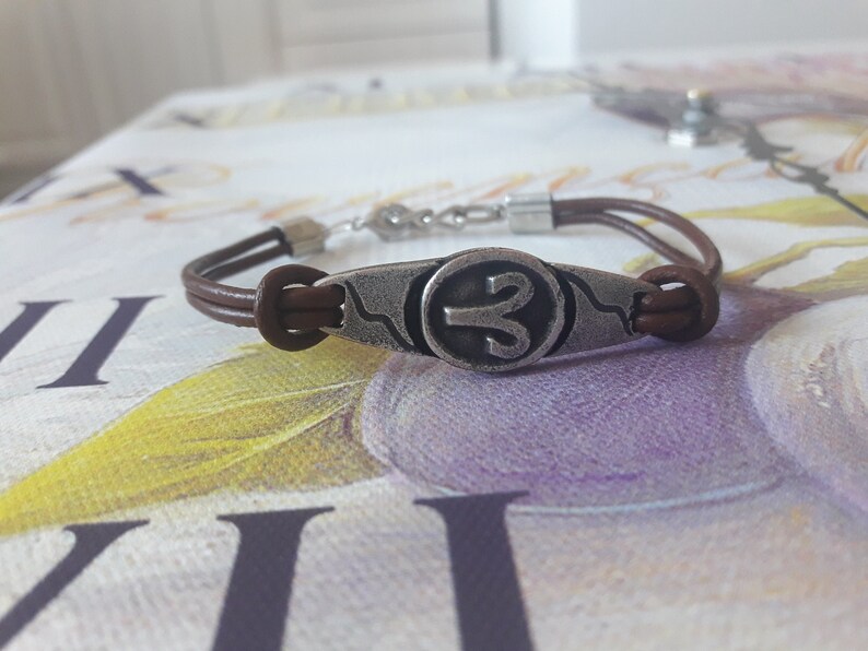 May include: A brown leather bracelet with a silver charm featuring the astrological symbol for Aries.