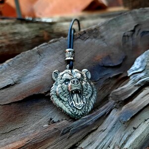 May include: A silver bear head pendant with a black leather cord. The bear's mouth is open and its teeth are visible. The pendant is on a wooden surface.