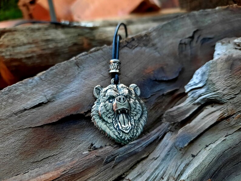 May include: A silver bear head pendant with a black leather cord. The bear's mouth is open and its teeth are visible. The pendant is on a wooden surface.