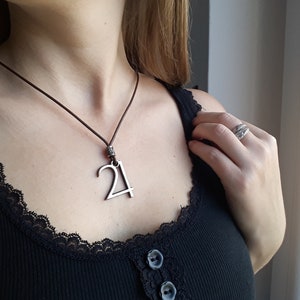 Puede incluir: Un collar de cuero marrón con un colgante de plata con forma de número 21.