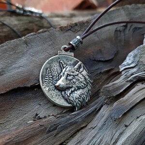 Op de afbeelding: Een zilveren ketting met een hanger in de vorm van een wolfskop. De wolf kijkt naar rechts en heeft een gedetailleerde vachtstructuur. De hanger is rond en heeft een boslandschap op de achtergrond. De ketting is gemaakt van een bruin leren koord.