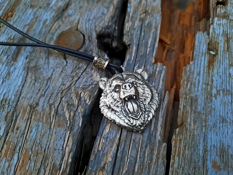 May include: A silver bear head pendant necklace with a black leather cord. The bear's mouth is open and its teeth are visible. The pendant is on a wooden surface.