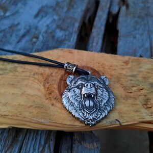 May include: A silver bear head pendant necklace with a black leather cord. The bear's mouth is open and its teeth are visible. The pendant is on a wooden surface.