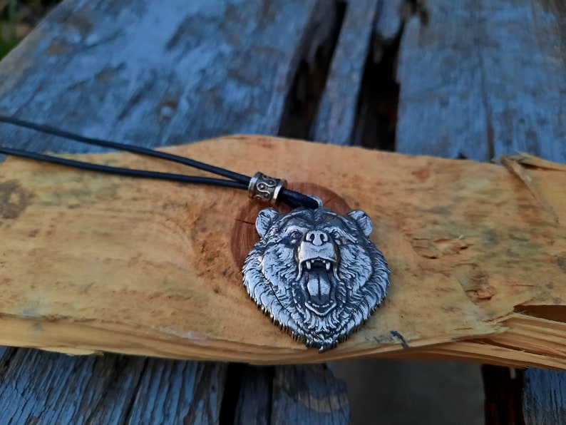 May include: A silver bear head pendant necklace with a black leather cord. The bear's mouth is open and its teeth are visible. The pendant is on a wooden surface.