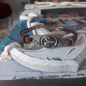 May include: A brown leather bracelet with a silver charm featuring the astrological symbol for Aries. The bracelet is displayed on a white and blue wooden anchor.
