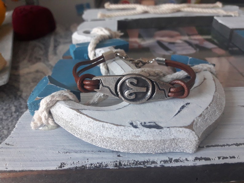 May include: A brown leather bracelet with a silver charm featuring the astrological symbol for Aries. The bracelet is displayed on a white and blue wooden anchor.
