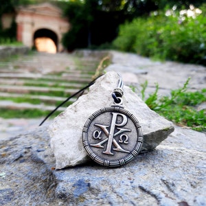 May include: A silver pendant necklace with a circular design featuring the Greek letters alpha and omega, and the symbol Chi Rho, on a rock with a blurred background of a stone pathway and green foliage.
