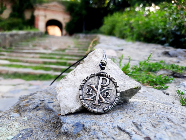 May include: A silver pendant necklace with a circular design featuring the Greek letters alpha and omega, and the symbol Chi Rho, on a rock with a blurred background of a stone pathway and green foliage.