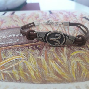 May include: A brown leather bracelet with a silver charm featuring the astrological symbol for Aries.