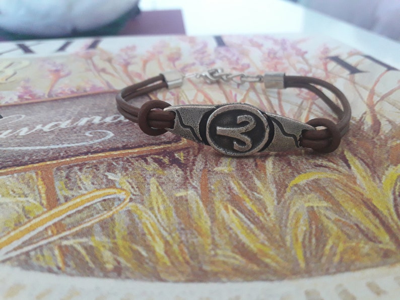 May include: A brown leather bracelet with a silver charm featuring the astrological symbol for Aries.
