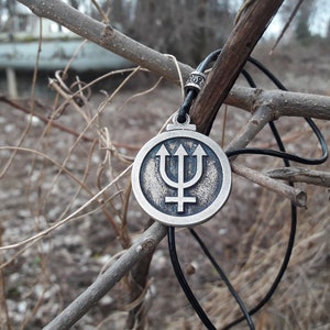 May include: A silver pendant necklace with a circular design featuring a trident and a symbol. The necklace is hanging from a branch with a black cord.