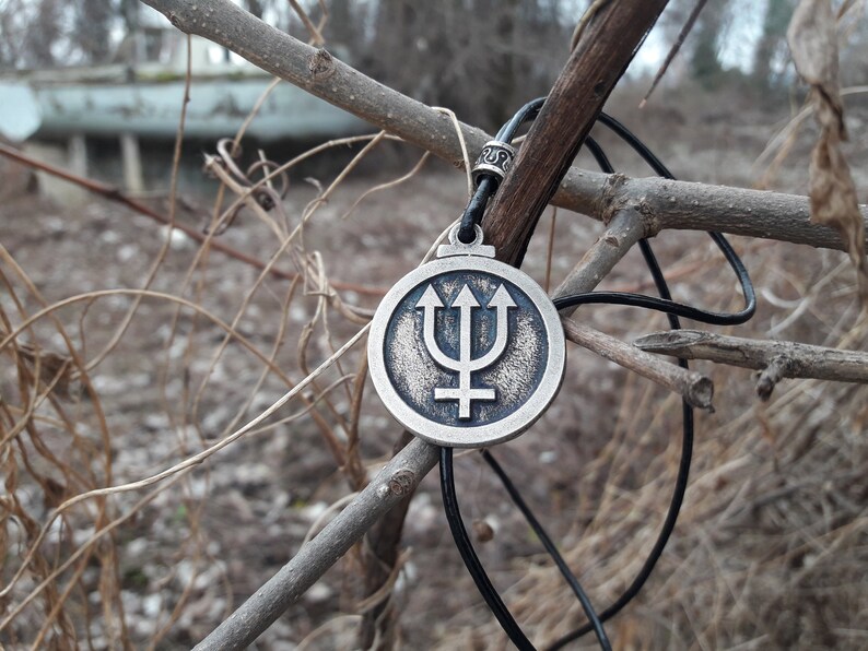 May include: A silver pendant necklace with a circular design featuring a trident and a symbol. The necklace is hanging from a branch with a black cord.