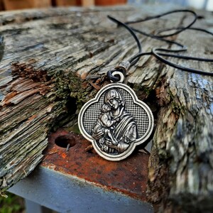 May include: A silver pendant necklace with a detailed image of the Virgin Mary holding a baby Jesus. The pendant is on a black cord and is resting on a weathered wooden surface.