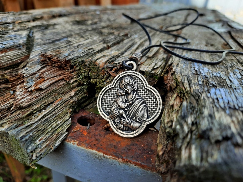May include: A silver pendant necklace with a detailed image of the Virgin Mary holding a baby Jesus. The pendant is on a black cord and is resting on a weathered wooden surface.